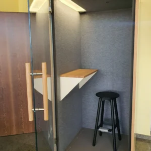 Schallisolierte Telefonkabine fürs Büro mit Glastür, Holzregal, grauer Akustikverkleidung und schwarzem Hocker